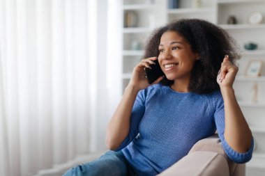Evde cep telefonuyla gülümseyen mutlu Afrikalı Amerikalı kadın, oturma odasında koltukta otururken cep telefonuyla eğlenen neşeli siyah kadın,