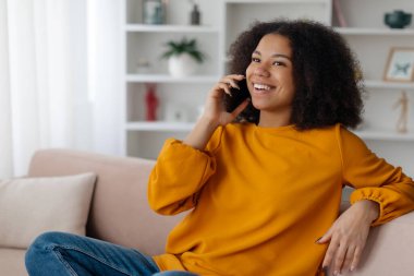 Kıvırcık saçlı genç bir Afrikalı Amerikalı kadın koltukta rahat bir şekilde oturur ve neşeli bir telefon görüşmesi yapar. Parlak, davetkar iç mekan rahatlatıcı bir atmosferi yansıtıyor..