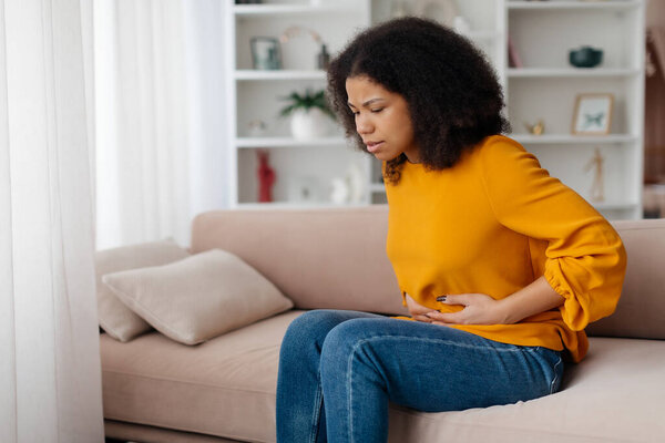 Живот Ache. Больной черный женщина страдает от острой боли в животе дома, расстроен афроамериканская женщина, имеющие менструальные боли или проблемы с пищеварением, сидя на диване и трогая живот