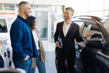 Satış Merkezinde yeni araba seçerken mutlu genç çift ile konuşuyor, Gülümseyen Milenyum Eşleri Modern Otomobil Showroom 'da Aile Aracı satın alıyor, Otomobil Gösterisi Yöneticisi