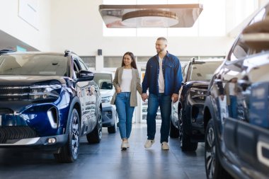 Genç bir çift modern araba galerisinde el ele yürüyor, mutlu eşler lüks araçları lüks arabalarla dolu lüks galerilerde satın almaya hazırlar.