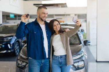 Yeni araba aldıktan sonra galeride selfie çeken mutlu çift, yeni arabalarını satın almalarını kutlayan neşeli eşler, akıllı telefon tutan kadın, gururla araba anahtarlarını gösteren adam.