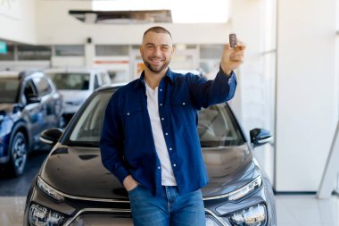 Otomobilin anahtarını elinde tutan yakışıklı bir adam, galeriden yepyeni bir araba alıyor. Mutlu milenyum erkeği yeni bir araba alıyor, siyah arabaya yaslanıyor ve anahtarı kameraya gösteriyor, boş alan.