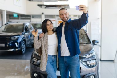 Yeni arabalarını aldıktan sonra modern araba galerisinde selfie çeken neşeli genç çift. Kadın gururla arabanın anahtarlarını tutarken, erkek akıllı telefonu tutuyor, neşeli anı yakalıyor.