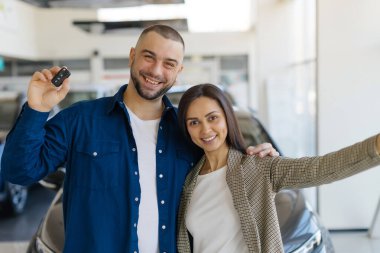 Yeni arabalarını aldıktan sonra modern araba galerisinde kutlama selfie 'si için poz veren neşeli genç çift. Kadın kolunu uzatıp ateş ederken adam gururla arabanın anahtarlarını tutuyor.