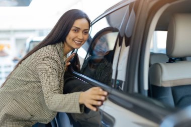 Modern galeride yeni arabasını heyecanla kucaklayan mutlu bir kadın. Arabanın parlak yüzeyi onun mutlu ifadesini ve yeni arabasıyla güçlü duygusal bağlantısını yansıtıyor..