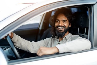 Beyaz bir arabada oturan gülümseyen Hintli adam, kolları pencerede dinleniyor, yakışıklı doğu erkek sürücü kameraya güvenle poz veriyor, sürüşün keyfini çıkarıyor, yakın plan