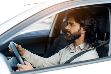 Sakince araba kullanan Hintli bir adam, pencereden dışarı bakıyor, odaklanmış ve huzurlu. Genç doğulu bir adam sabah ofisine gidiyor, aracını sürüyor, lüks bir otomobilin içinde oturuyor, yandan bakıyor.