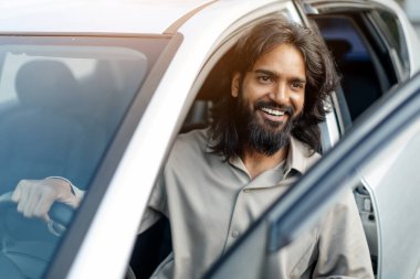 Mutlu Hintli adam arabanın içinde oturuyor, açık kapıdan gülümsüyor, yakışıklı milenyum doğu erkeği direksiyonu tutuyor ve kameraya bakıyor, yakın çekim.