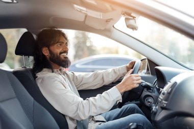 Gülümseyen Hintli adam araba sürüyor ve gülümsüyor, sürücü koltuğunda oturuyor ileriye bakıyor, rahatlamış doğu erkeği iki eli direksiyonda, yan görüşlü sürüşün tadını çıkarıyor.