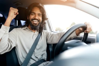 Arabada gülümseyen Hintli bir adam, araba sürerken yumruğunu kaldırarak kutluyor, bej gömlek ve emniyet kemeri takmış, kameraya gülümsüyor, gezinin keyfini çıkarıyor.