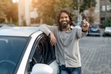 Bir şehir caddesinde beyaz arabasının yanında duran gülümseyen Hintli adam, gururla arabanın anahtarlarını kameraya gösteriyor, neşeli doğu erkeği yeni bir araba satın alıp, sürmekten zevk alıyor.