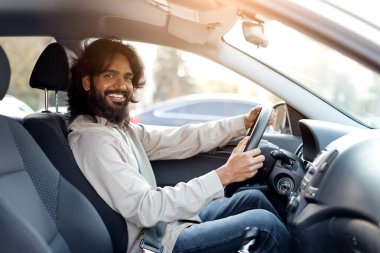 Arabadaki gülümseyen Hintli adam, yakışıklı doğu erkeği kameraya bakarken rastgele poz veriyor, sürücü ellerini direksiyonda tutuyor, pencereden içeri güneş ışığı giriyor.