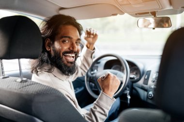 Sürücü koltuğundaki heyecanlı Hintli adam, arkasını dönüyor ve sevinçle sevinçle yumruklarını kaldırarak kutluyor, mutlu, aşırı neşeli doğu erkeği yeni araba almaya duygusal tepki veriyor, araba sürmekten zevk alıyor.