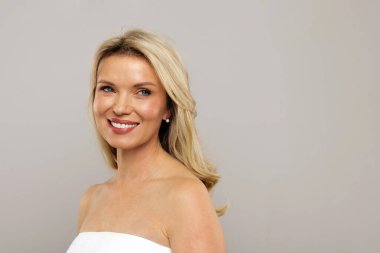 Orta yaşlı güzel bir kadının portresi zarif bir şekilde gülümseyen, çekici olgun bir kadın açık gri stüdyo arka planına sarılıp havluya sarmalanmış, fotokopi çekiyor.