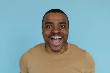 Neşeli genç siyahi adamın portresi heyecanla ağzını açıyor ve kameraya bakıyor, bej tişört giyen Afrikalı Amerikalı erkek açık mavi arka planda duruyor, boşluğu kopyalıyor.