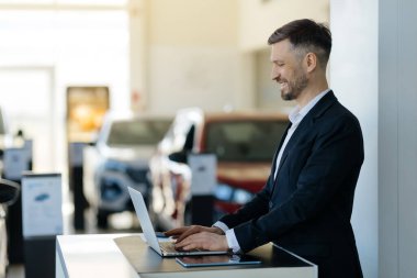 Parlak modern araba galerisinde dizüstü bilgisayar kullanarak resmi kıyafet giymiş gülümseyen bir satıcı, oto kuaförde masasında duran ve bilgisayarda yazı yazan yakışıklı genç bir adam, fotokopi uzayı ile yan görüş görüntüsü.