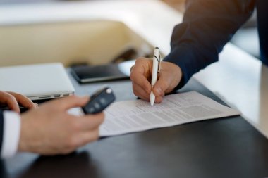 Müşteri sözleşmesi imzalarken yakın plan bir galeri merkezinde araba anahtarlarını tutan satış elemanı, araba galerisinde araba alan tanınmayan bir adam, belgeleri dolduran,