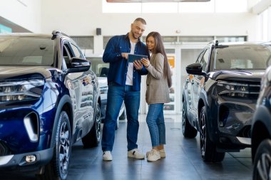 Genç mutlu çift, modern bayilik galerisinde dururken dijital tablette araba özelliklerini kontrol ediyor. Gülümsüyor, milenyum erkeği ve kadını yeni aile aracını seçiyor.