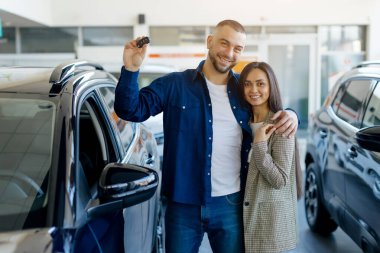 Gülümseyen milenyum çifti araba anahtarlarını gururla tutarken, modern galeride yeni bir aracın yanında dururken, mutlu genç eşler yeni araba alıyor, alışverişi kutluyor.