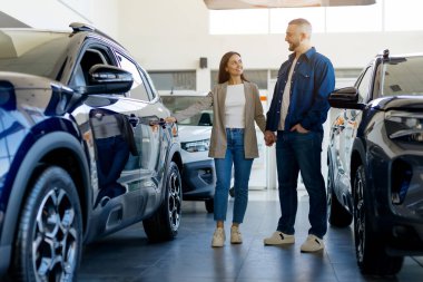 Gülümseyen genç bir çift el ele tutuşuyor, parlak ve modern bir galeride arabaları inceliyor, kadın arabanın kapı koluna dokunuyor, mutlu aile yeni bir araç seçiyor.