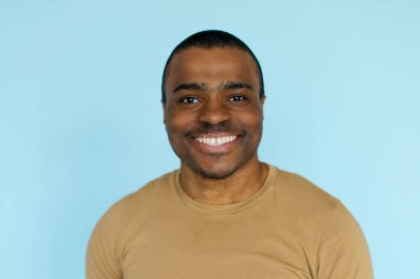 Afro-Amerikalı bir adamın neşeli portresi doğrudan kameraya gülümsüyor, gündelik bronz gömlekli genç bir erkek mavi bir stüdyo arka planında duruyor, fotokopi alanı ile yakın çekim.