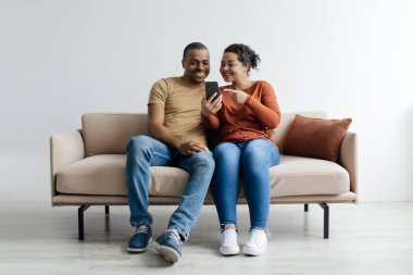 Güzel siyah çift cep telefonlarını evde birlikte kullanıyor, mutlu Afrikalı Amerikalı eşler modern akıllı telefonlarıyla koltukta dinleniyor, yeni uygulama veya alışveriş sitelerine bakıyor,