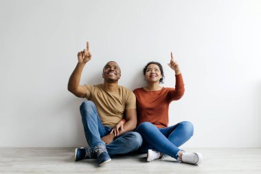 Kafalarının üstündeki fotokopi boşluğunu işaret eden mutlu siyah çift, yerde oturan ve beyaz arka planda bedava yer gösteren Afro-Amerikan erkek ve kadın teklif ya da reklam için