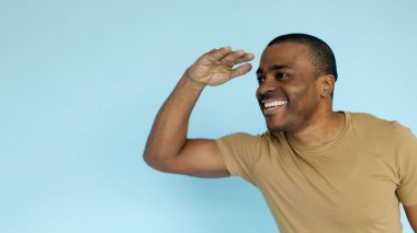 Bronz tişörtlü, alnına yakın bir şekilde gülümseyen ve sanki mesafeyi arıyormuş gibi şakacı hareketler yapan Afro-Amerikan bir adam, mavi stüdyo arka planında, panorama