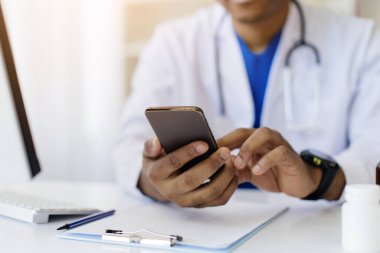 Afro-Amerikan doktorların elleri kapalı, akıllı telefon kullanıyorlar, tanınmayan siyah erkek terapist masasında oturuyor, modern kliniğin iç kısımlarında klavye ve klavye ile kesilmiş.