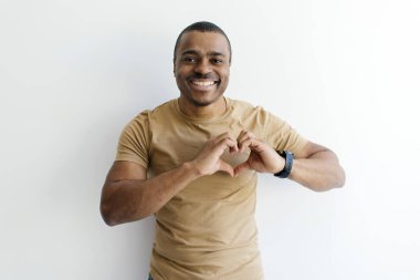 Gülümseyen genç siyah adam, elleri göğsüne yakın bir şekilde kalp hareketi yapıyor, beyaz stüdyo arka planında poz verirken, mutlu Afrikalı Amerikalı adam kamerada sevişiyor, boşluğu kopyalıyor.