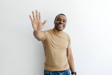 Dost canlısı siyah adam elini kaldırıp kameraya gülümsüyor ve merhaba diyor. Beyaz stüdyo arka planında poz veren neşeli, Afro-Amerikan erkek.