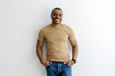 Bronz tişörtlü siyahi genç adam, elleri ceplerinde güvenle gülümsüyor, mutlu Afro-Amerikan erkek, beyaz bir stüdyonun arka planında duruyor ve kameraya bakıyor, boşluğu kopyalıyor.
