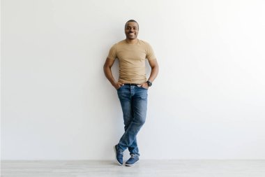 Gülümseyen Afro-Amerikan bir adamın, elleri cebinde, beyaz bir duvara yaslanmış, bronz bir tişört ve kot pantolon giyen yakışıklı siyahi bir adamın, fotokopi çekimleri.