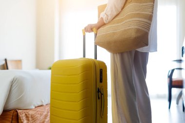 Otel odasında parlak sarı bir bavul ve örülmüş bir plaj çantasıyla duran kadının yakın görüntüsü. Oda sıcak bir şekilde ışıklandırılmış ve ayarı seyahat ya da tatil izlenimi uyandırıyor.