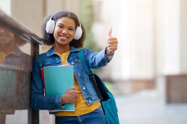 Güzel Siyah Kız Öğrenci Açık havada üniversite binasının yanında duruyor, gülümseyen genç kadın çalışma kitaplarını tutuyor ve kablosuz kulaklık takıyor, uzay kopyası yapıyor.