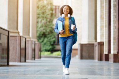 Üniversite kampüsünde, elinde çalışma kitapları ve kahve fincanı, kulaklık ve kot ceket giyen gülümseyen siyah bir kadın.
