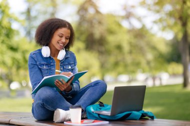 Genç Afrikalı Amerikalı kız öğrenci dışarıda sınava hazırlanıyor, laptoplu ve çalışma kitaplarıyla gülümseyen siyah kadın kampüsün dışında çalışıyor, bankta oturuyor, boşluğu kopyalıyor.