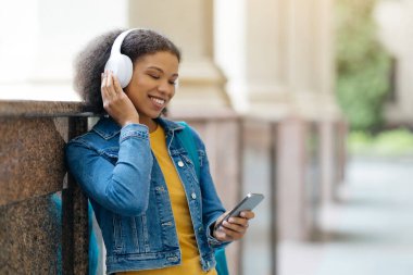 Siyahi Afrikalı Amerikalı kız öğrenci akıllı telefondan müzik dinliyor. Kablosuz kulaklıkla gülümsüyor. Neşeli genç bir kadın kampüste dinlenirken favori çalma listesinin keyfini çıkarıyor.