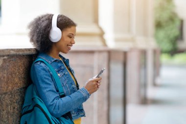 Gülümseyen siyah kız öğrenci müzik dinliyor ve akıllı telefonlarda internette geziniyor kampüste dinlenirken, kulaklık takan mutlu Afro-Amerikan kadın favori müzik listesinin keyfini çıkarıyor.