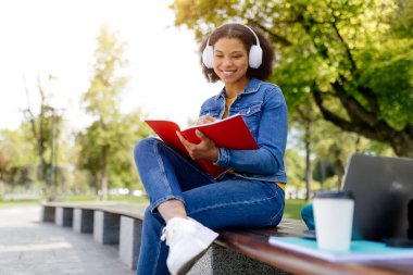 Afro-Amerikalı bir kız öğrenci parkta bir bankta bağdaş kurarak oturuyor, elinde kırmızı bir defter, dışarıda çalışırken gülümsüyor, kulaklıkları ve kot ceketi ile, bedava uzay boşluğu.