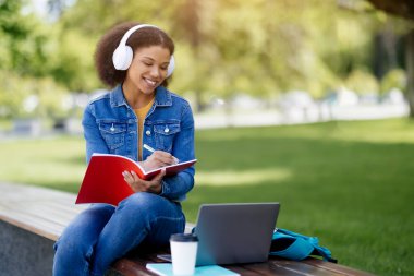 Genç Afrikalı Amerikalı kadın dışarıda dizüstü bilgisayar ve not defteriyle çalışıyor, dışarıda sınava hazırlanan gülümseyen siyah kız öğrenci, parkta bankta oturuyor, çevrimiçi eğitimin tadını çıkarıyor,