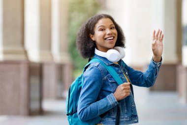 Afro-Amerikalı kız öğrenci kampüste yürürken mutlu bir şekilde el sallıyor. Pozitif siyah kadın kulaklık, kot ceket ve parlak sırt çantası takıyor. Neşe ve pozitifliği ifade ediyor.