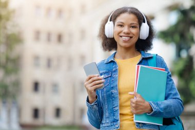 Modern öğrenci. Çalışma kitaplarıyla gülümseyen siyah bir kadının portresi ve el telefonuyla dışarıda poz veren, sırt çantalı ve kablosuz kulaklıklı güzel bir Afro-Amerikan kadını.