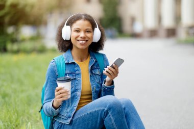 Siyah kız öğrenci dışarıda akıllı telefon ve kahveyle rahatlıyor, mutlu Afro-Amerikan kadın çimlerin üzerinde oturuyor, cep telefonu ve fincanı tutuyor, kampüste boş vakitlerin tadını çıkarıyor.