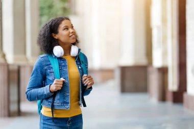 Afrika kökenli Amerikalı bir kız öğrenci, üniversite kampüsünde yürürken, kulaklık, kot ceket ve sırt çantası takan, gülümseyen siyah bir kadın.