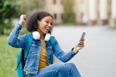 Siyah kız öğrenci elinde akıllı telefonuyla kaldırımda otururken sevinçle kutluyor. Afro-Amerikan kadın cep telefonuna bakıyor, kulaklık takıyor ve kot ceket giyiyor.