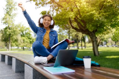 Afro-Amerikalı kız öğrenci dışarıda dizüstü bilgisayarla oturuyor ve başarıyı kutluyor, neşeli siyah kadın sevinçle yumruk kaldırıyor, kampüs parkında çalışırken gülümsüyor, yer kopyalıyor.
