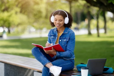 Parktaki bankta oturan Afro-Amerikalı bir kız öğrenci, kırmızı defterine yazıyor, kulaklık takan ve kot ceket giyen gülümseyen siyah bir kadın, açık havada çalışırken güneşli bir günün tadını çıkarıyor.