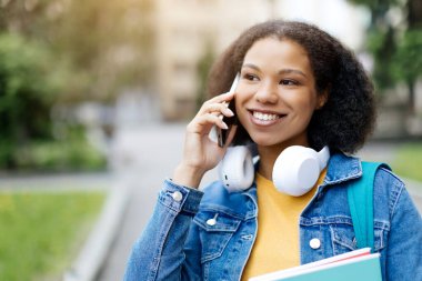 Dışarıda yürürken cep telefonuyla konuşan mutlu Afrikalı Amerikalı öğrenci, çalışma kitaplarını tutan neşeli siyah kadın ve arkadaşınla keyifli telefon konuşmaları, kopyala.